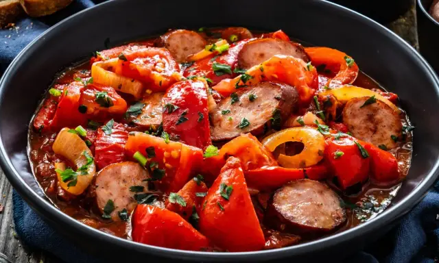 Pyszny obiad z kiełbasą, papryką i pomidorami w sosie, posypany natką pietruszki. Obok kromki chleba.