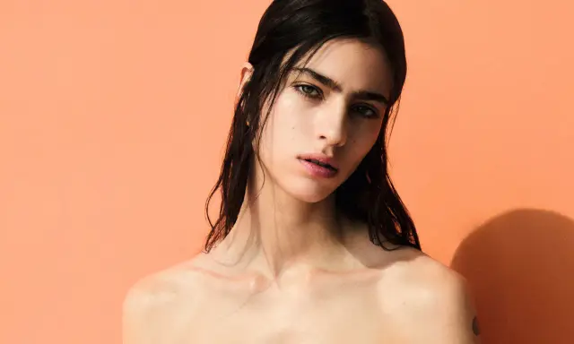 A striking portrait of Loli Bahia, her dark hair cascading around her shoulders against a vibrant peach backdrop.