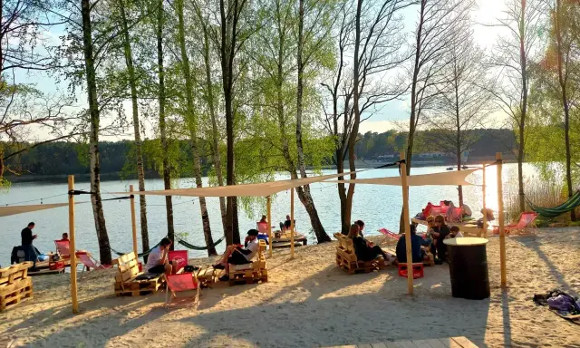 Pola namiotowe nad jeziorem, ludzie odpoczywają na plaży pod baldachimami, wśród drzew.