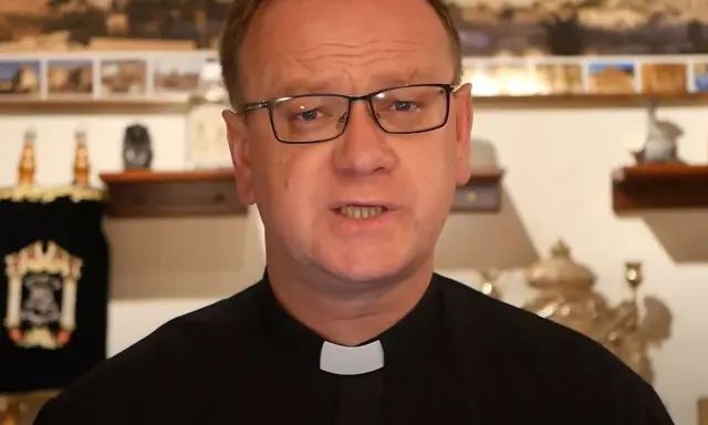 Wojciech Węgrzyniak w okularach, w czarnej sutannie z białą kolaretką, przemawia. W tle widać zwoje Tory i obrazy.