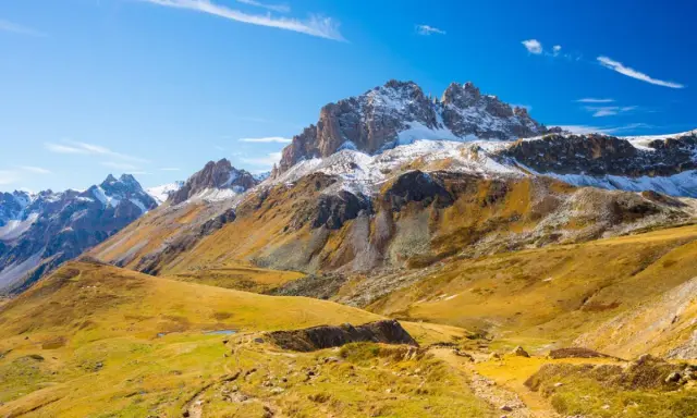 Ampia **foto grandangolo** di montagne innevate e pendii dorati autunnali, con un sentiero che serpeggia tra l'erba.