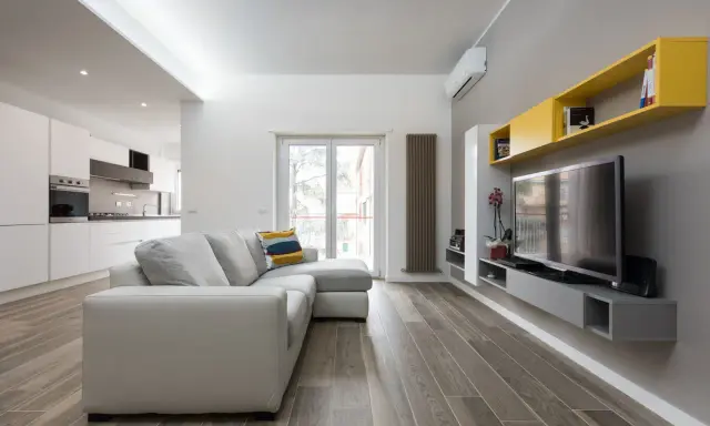 Un progetto soggiorno stretto e lungo con divano grigio, TV a parete e mensole gialle. La cucina moderna è visibile sullo sfondo.