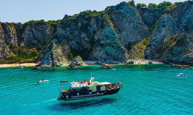 Barca turistica naviga nelle acque cristalline vicino al porto di Tropea, con imponenti scogliere a picco sul mare e piccole spiagge affollate.