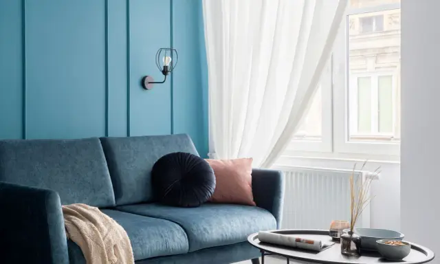 Divano blu petrolio con cuscini decorativi e coperta. Parete azzurra con applique. Abbinamenti moderni e rilassanti.