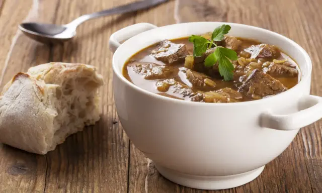 Aromaticzny gulasz wołowy w białej miseczce, ozdobiony natką pietruszki, obok kromka chleba i łyżka.