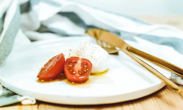 Kuleczki mozzarelli z pomidorkami koktajlowymi na białym talerzu, obok złote sztućce i ściereczka.
