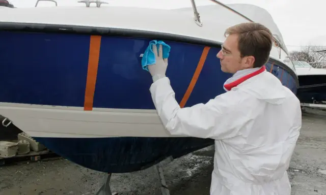 Un homme en combinaison blanche effectue l'entretien d'un bateau bleu et blanc, le lustrant avec un chiffon bleu.