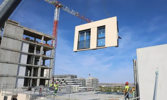 Grúa elevando un módulo prefabricado para la construcción industrializada de un edificio. Obreros supervisan la operación.