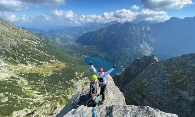 Dwóch wspinaczy na szczycie, z widokiem na Tatry i Morskie Oko. W tle góry i jezioro.