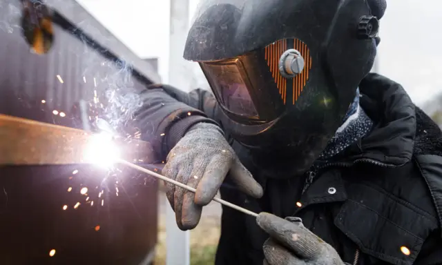 Soldador con casco y guantes usa un electrodo para unir metales, creando chispas brillantes y humo.