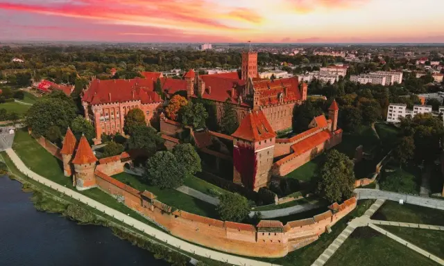 Zamek krzyżacki w Malborku, imponująca twierdza z czerwonej cegły, otoczona fosą i zielenią, pod rozgwieżdżonym, różowym niebem. Ciekawostki o jego historii czekają na odkrycie.