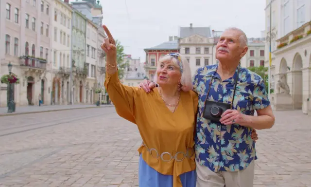 Starsze małżeństwo odkrywa urokliwe, przyjazne seniorom miasta. Kobieta z aparatem wskazuje na zabytkową architekturę.
