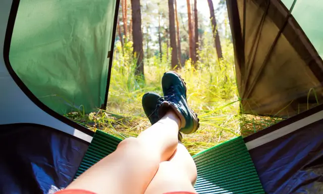 Nogi w butach trekkingowych odpoczywają w namiocie. Słońce przebija się przez drzewa. Idealne miejsce, by zanocuj w lesie.