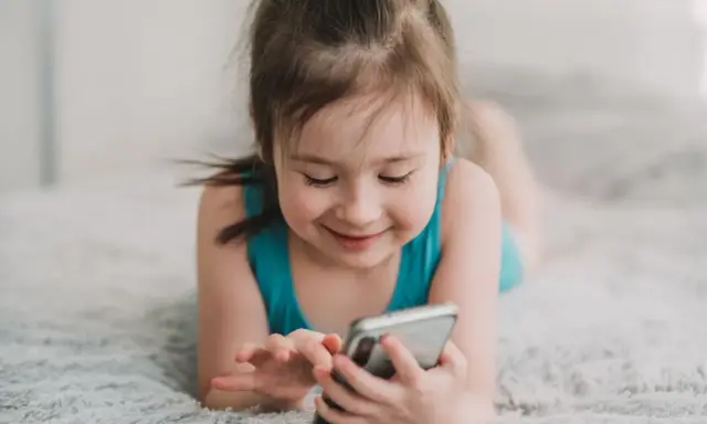 Uśmiechnięta dziewczynka leży na dywanie, bawiąc się telefonem. Wpływ telefonu komórkowego na mózg dziecka jest tematem dyskusji.