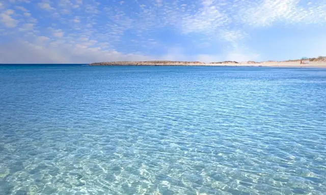 Crystal clear turquoise water with gentle ripples, leading to a sandy beach and distant dunes under a blue sky with scattered clouds.
