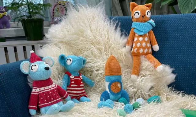 En virkad amigurumi katt med orange och vita prickar sitter på en lurvig matta bredvid en blå mus med tomteluva och en blå elefant i hängselbyxor.
