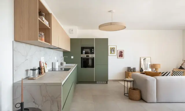 Cuisine moderne avec des armoires vert olive, un plan de travail en marbre et des étagères en bois clair. Un salon adjacent est visible.