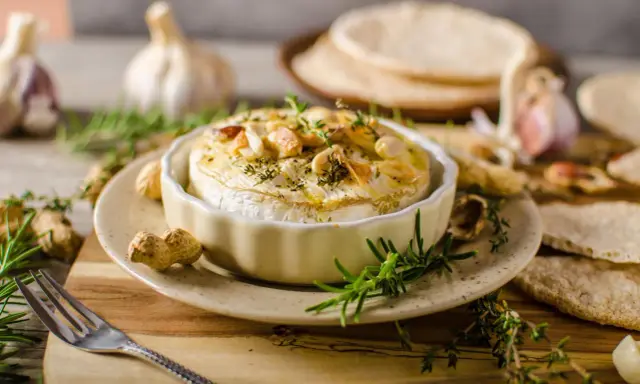 Pieczony camembert w naczyniu, ozdobiony ziołami i orzechami, z czosnkiem i pieczywem w tle.