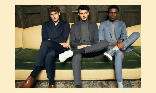 Three men in suits sit on a couch. The man on the left wears dark dress pants, while the other two wear suit pants.