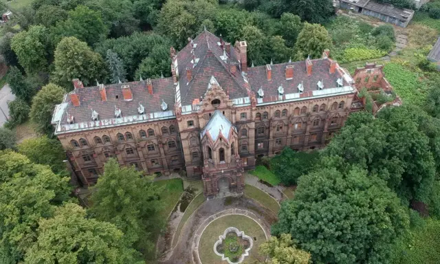 Mokrzeszów pałac, rozległy, ceglany budynek z charakterystycznym dachem, otoczony bujną zielenią.