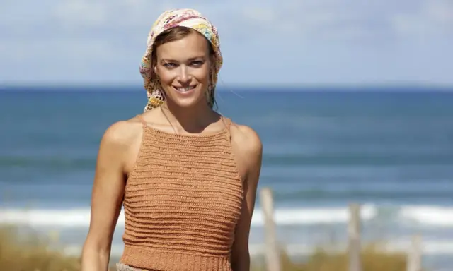 Woman in a crocheted top and headscarf smiles at the camera on a beach.