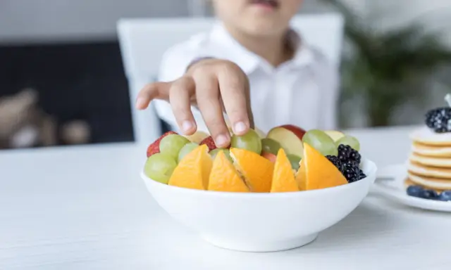 Małe rączki sięgają po miskę pełną owoców: pomarańczy, truskawek, jabłek i winogron dla niemowlaka.