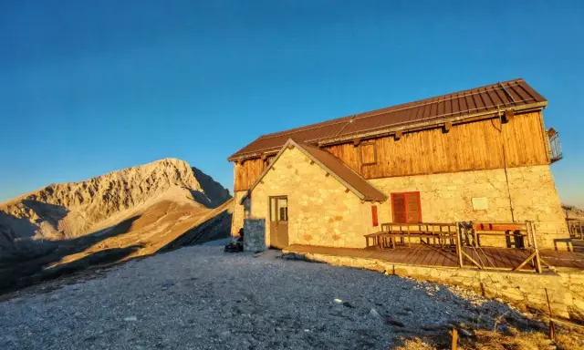 Il rifugio Duca degli Abruzzi, con la sua struttura in pietra e legno, si erge maestoso al tramonto, dominando un paesaggio montano aspro e solitario.