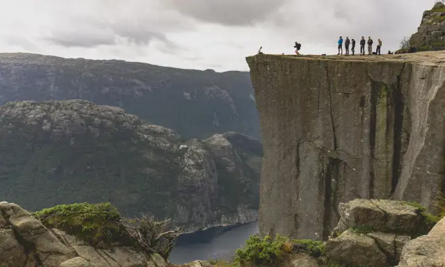 Grupa ludzi na krawędzi klifu w **Stavanger, Norwegia**, z widokiem na fiord i góry.