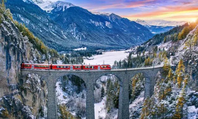 Il trenino del Bernina attraversa un viadotto in pietra in un paesaggio alpino innevato, con montagne maestose e un cielo colorato al tramonto.