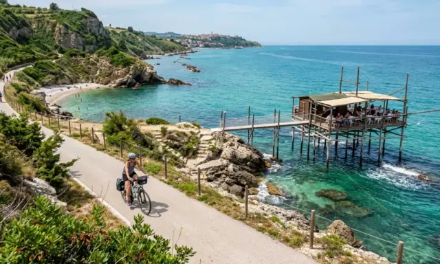 Ciclista lungo la via dei trabocchi, con un trabocco in legno sull'acqua cristallina e una spiaggia rocciosa.