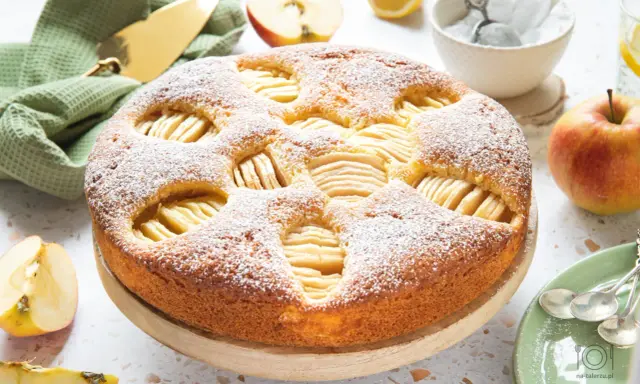 Szybkie tanie ciasto z jabłkami, posypane cukrem pudrem, prezentuje się apetycznie. Obok jabłka, cytryna i zielony ręcznik.
