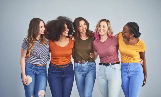 Cinque amiche sorridono, vestite con jeans e magliette colorate, tra cui spiccano tonalità marrone e bianco.