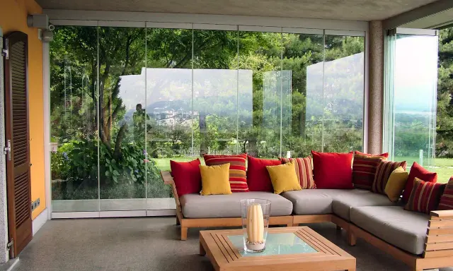 Come arredare una veranda aperta: un divano angolare grigio con cuscini rossi e gialli, un tavolino in legno e una vista sul giardino.