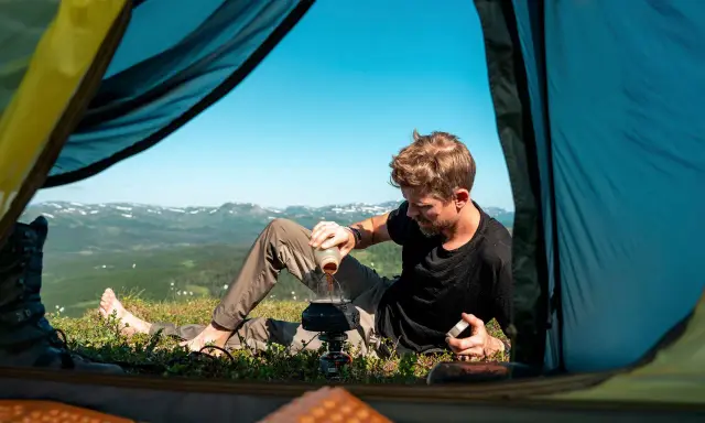 Man förbereder kaffe utanför tältet med en fantastisk utsikt. En del av packlista vandring sommar.