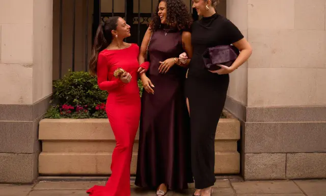 Three women in elegant gowns, discussing what not to wear to a wedding. One in red, one in plum, and one in black.
