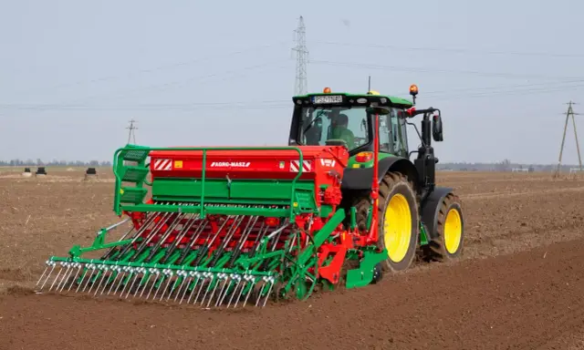 Zielony traktor z siewnikiem Agro-Masz pracuje na polu. Maszyna precyzyjnie wysiewa nasiona, optymalizując ile trawy na hektar zostanie zasiane.