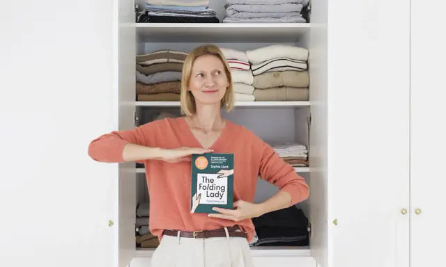 Kobieta z książką "The Folding Lady" pokazuje, jak składać koszulki, stojąc przed otwartą szafą pełną schludnie ułożonych ubrań.