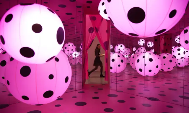 A woman walks through a mirrored room filled with pink polka-dotted spheres, a signature of Yayoi Kusama's immersive art.