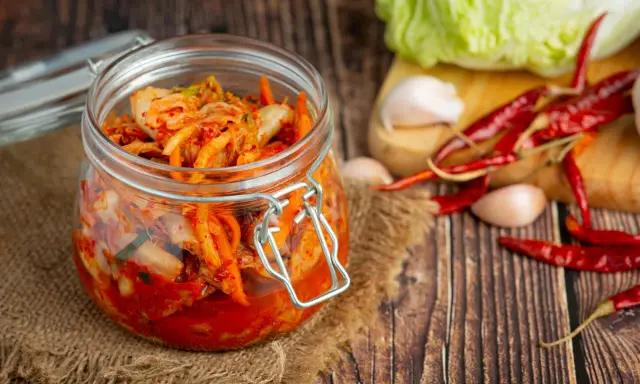 Słoik z kimchi na tle desek, obok kapusta, czosnek i papryczki chili.
