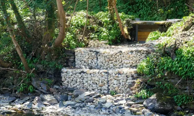 Estructura de muro de gavion con piedras blancas, rodeada de vegetación y rocas junto a un río.