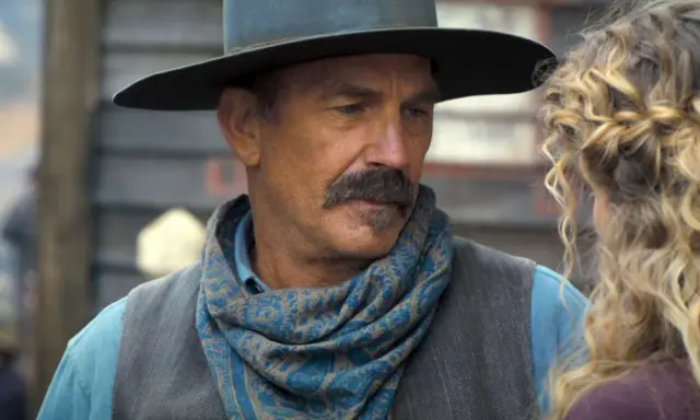 Kevin Costner w filmie z westernowym kapeluszem i chustą na szyi, rozmawia z kobietą.