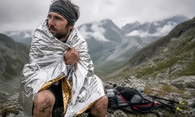 Un coureur transi, enveloppé dans une couverture de survie, contemple le paysage montagneux. Son sac à dos et ses bâtons de randonnée sont posés à côté.