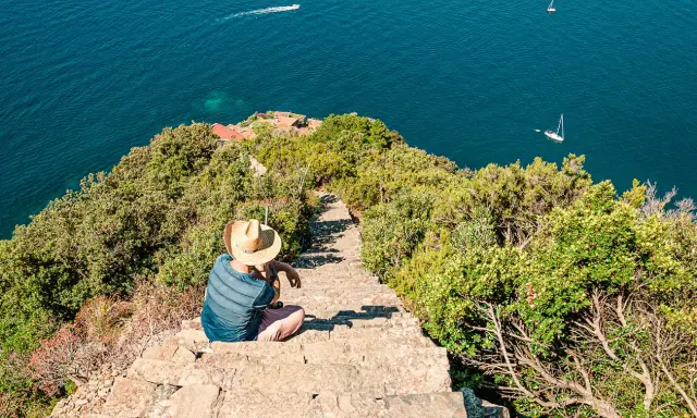 Un uomo con cappello di paglia seduto su una scalinata di pietra guarda il mare blu. Un sentiero scende verso il mare, suggerendo come arrivare a Monesteroli.