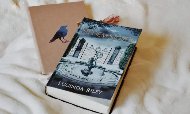Okładka książki "Siedem Sióstr" Lucindy Riley na białym tle. Obok leży brązowy notes z wizerunkiem kruka.
