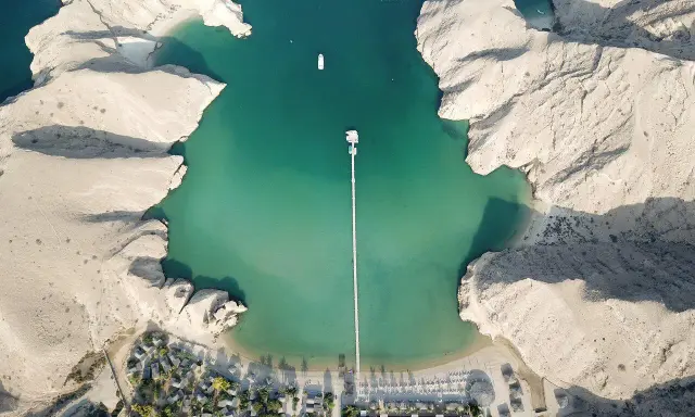 Vista aerea di un resort in Oman con bambini che giocano sulla spiaggia. Acque turchesi, montagne rocciose e un lungo pontile.