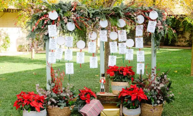 Decoración ceremonia civil con un arco de ramas y hojas, colgando tarjetas de invitados. Flores rojas y cestas adornan la base.