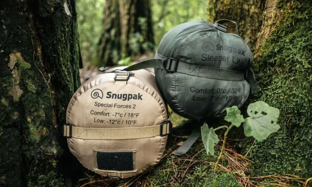 Dwa śpiwory Snugpak, jeden beżowy (komfort -7°C), drugi zielony (komfort 0°C), leżą na leśnym poszyciu.