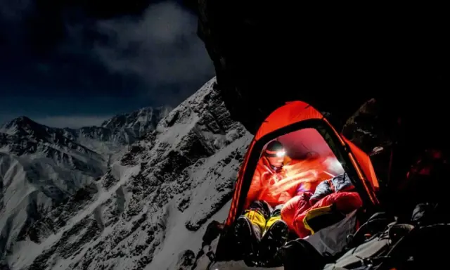 Nocny obóz na zboczu himalajskiego parbatu. W pomarańczowym namiocie rozświetlonym czołówką odpoczywa alpinista, otoczony surowym, ośnieżonym krajobrazem.