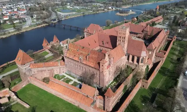 Potężny zamek krzyżacki, największy zamek w Europie, wznosi się nad rzeką Nogat. Czerwona cegła i liczne wieże tworzą imponujący widok.