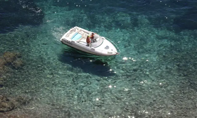 Deux personnes dans un bateau blanc, au moteur en panne, dérivent sur une eau turquoise et claire, révélant les fonds rocheux.